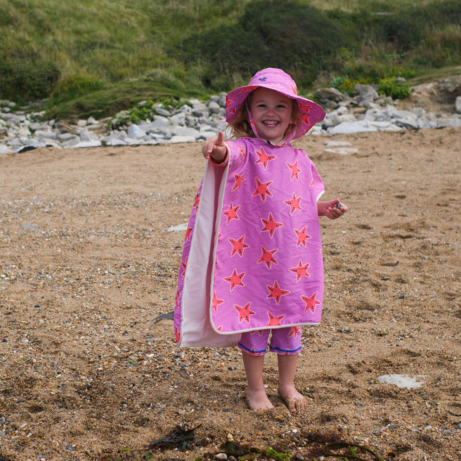 A happy child on a beach, wearing a UV Protective Sun Hat Pink Starfish, pointing towards the camera with a big smile.